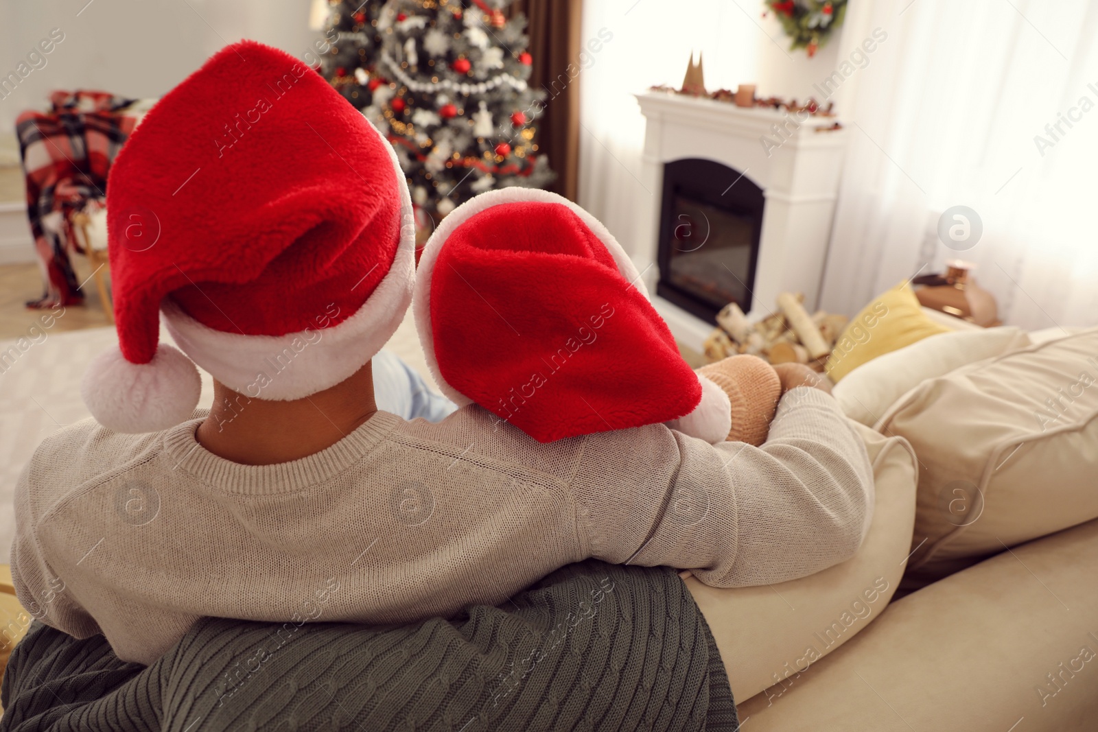 Photo of Couple in Santa hats sitting on sofa in living room decorated for Christmas, back view