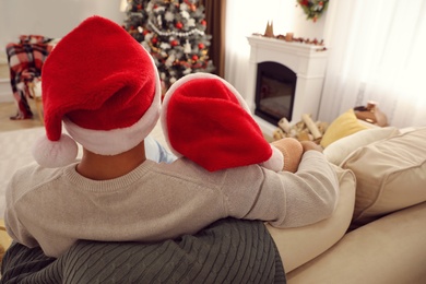 Photo of Couple in Santa hats sitting on sofa in living room decorated for Christmas, back view