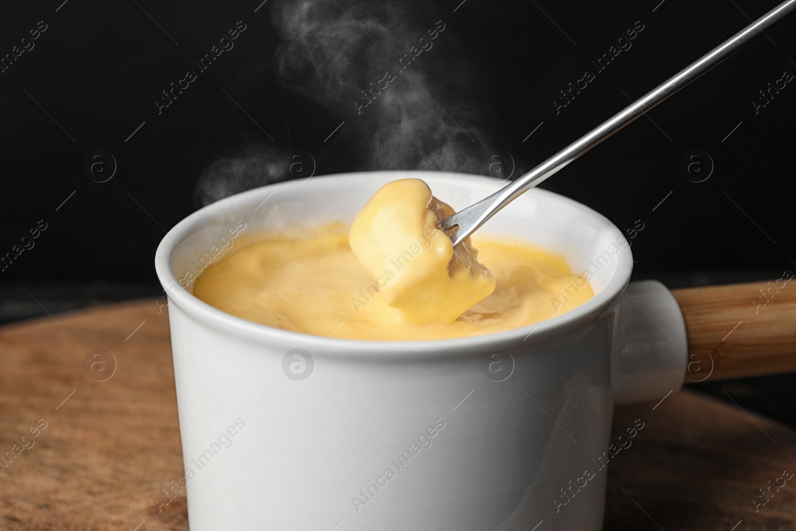 Image of Dipping piece of bread into fondue pot with tasty melted cheese at table, closeup