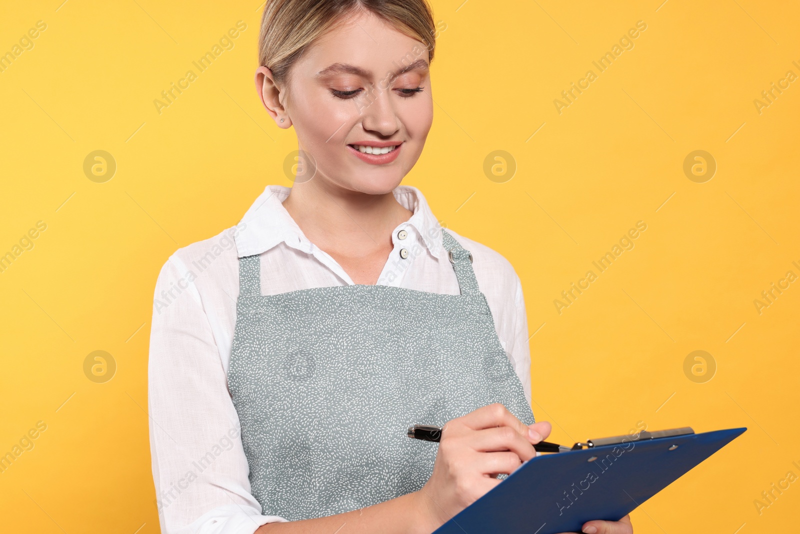 Photo of Beautiful young woman in clean apron with clipboard on orange background