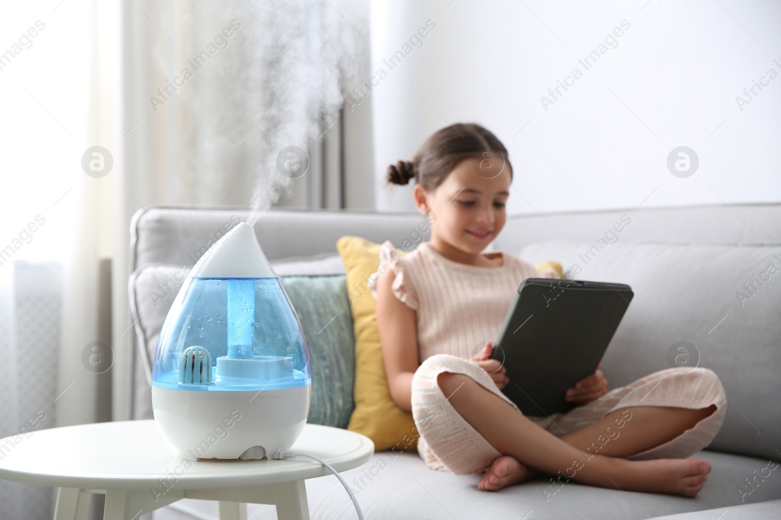 Photo of Little girl using tablet in room with modern air humidifier