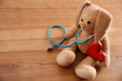 Photo of Toy bunny, stethoscope, heart and space for text on wooden background. Children's doctor