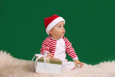 Photo of Cute baby in Santa hat with Christmas gift on fluffy carpet against green background