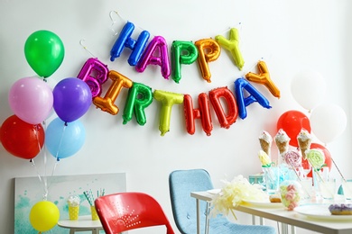 Table with treats and phrase HAPPY BIRTHDAY made of colorful balloon letters in living room