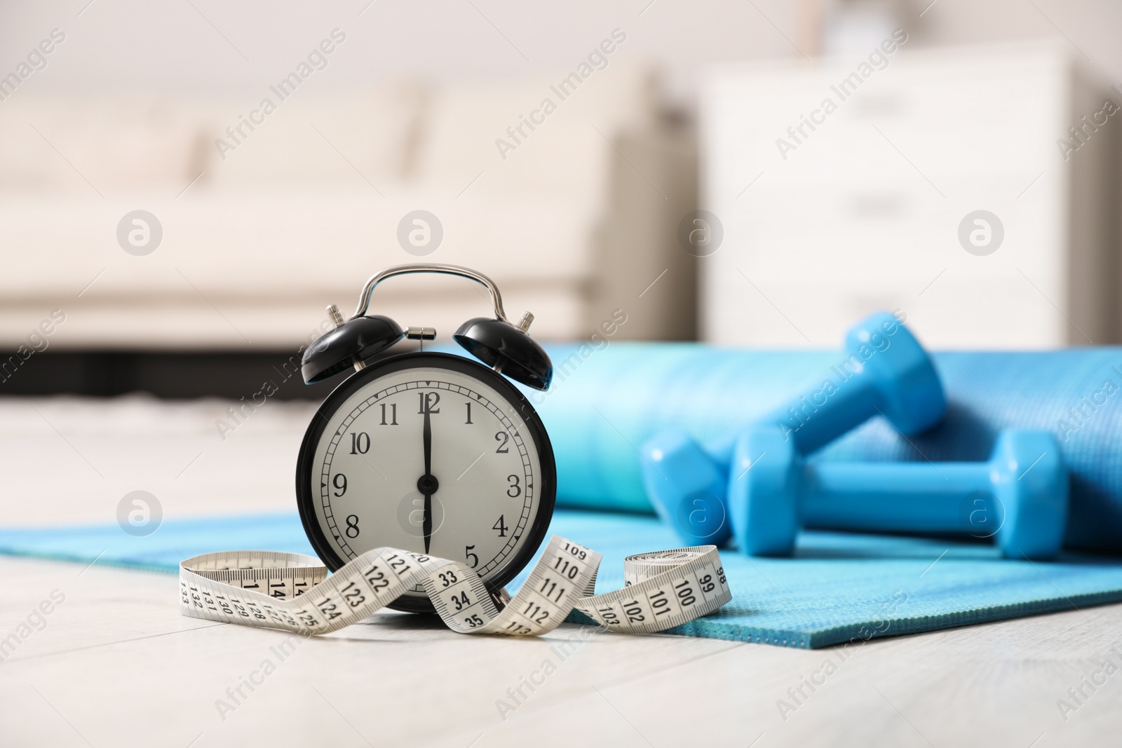 Photo of Alarm clock, yoga mat, measuring tape and dumbbells on wooden floor indoors. Morning exercise