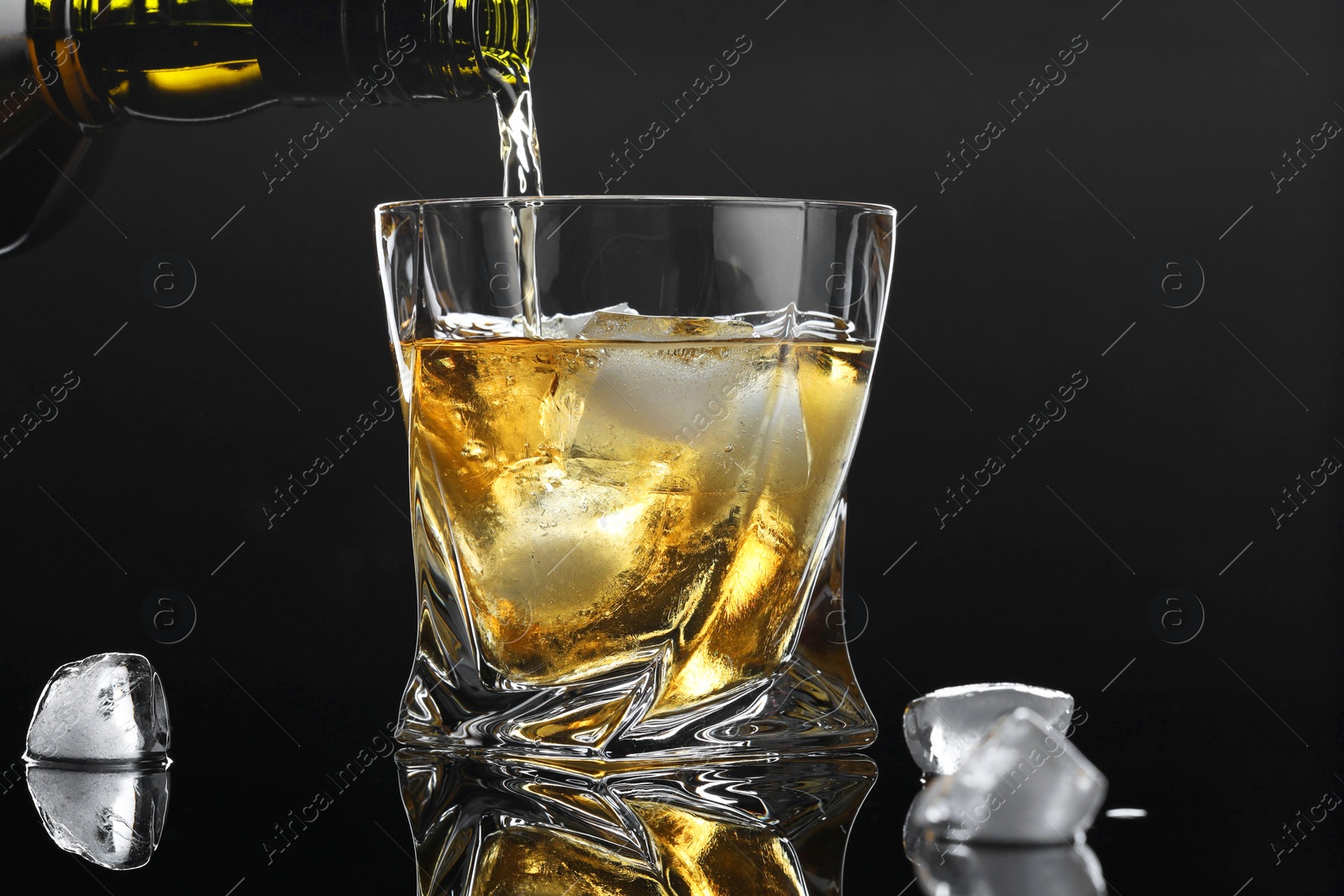 Photo of Pouring tasty whiskey from bottle into glass with ice at mirror table against black background, closeup