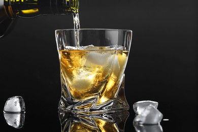 Pouring tasty whiskey from bottle into glass with ice at mirror table against black background, closeup