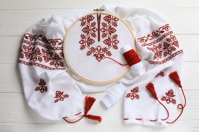 Shirt with red embroidery design in hoop, needle and threads on wooden table, flat lay. National Ukrainian clothes