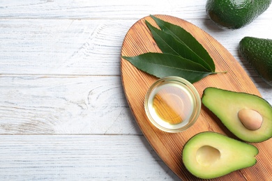 Board with bowl of natural oil and avocados on white wooden background, flat lay. Space for text