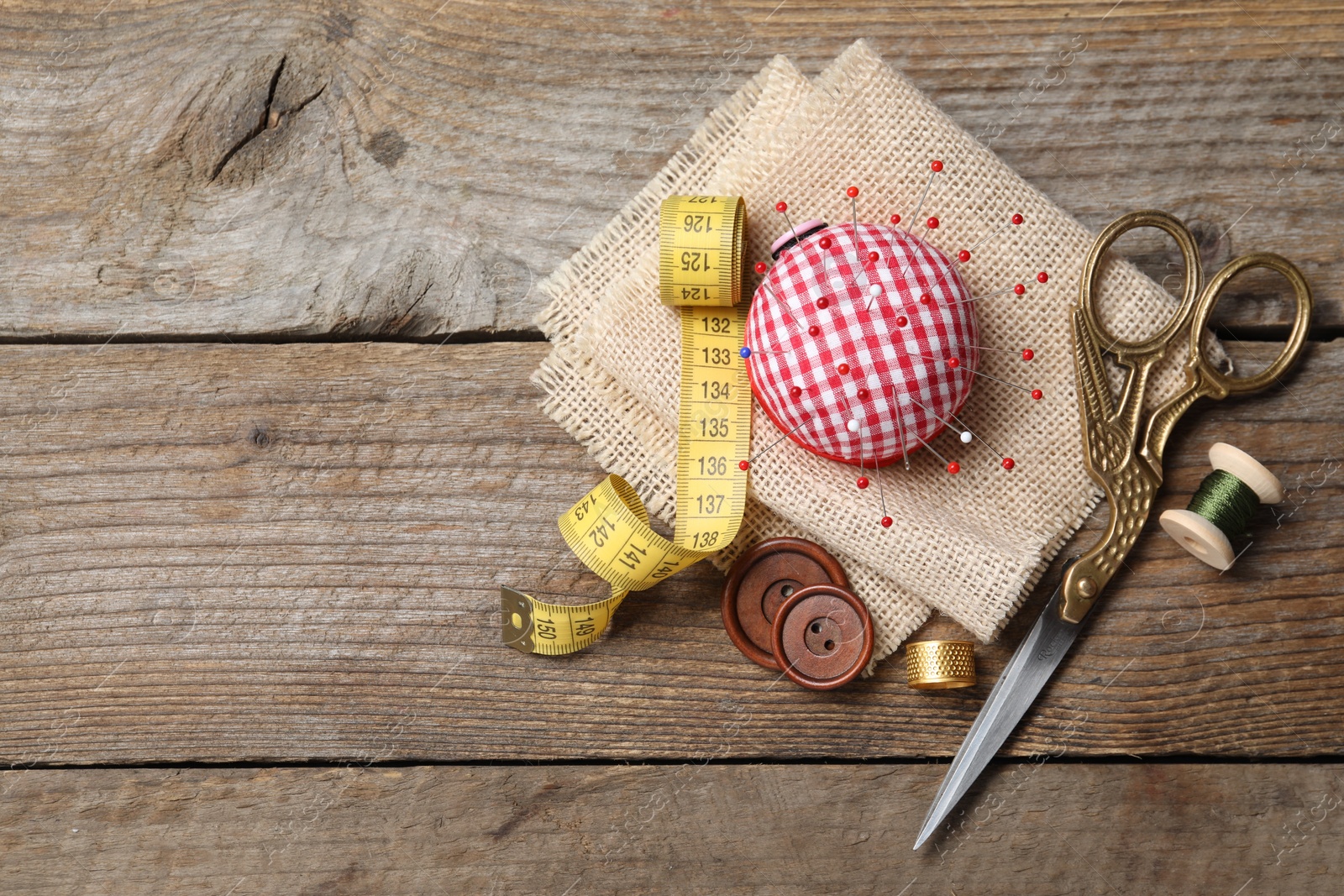 Photo of Checkered pincushion with pins and other sewing tools on wooden table, flat lay. Space for text