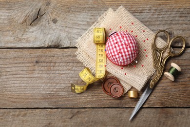 Checkered pincushion with pins and other sewing tools on wooden table, flat lay. Space for text