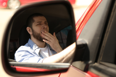 Photo of Tired man yawning in his auto, view through car side mirror