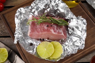 Photo of Aluminum foil with raw meat, rosemary, lime and spices on wooden table, above view