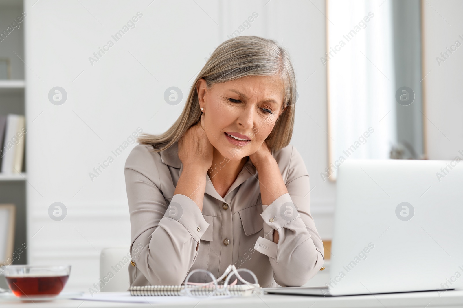 Photo of Woman suffering from neck pain at workplace in room