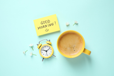 Photo of Delicious coffee, alarm clock and card with GOOD MORNING wish on light blue background, flat lay