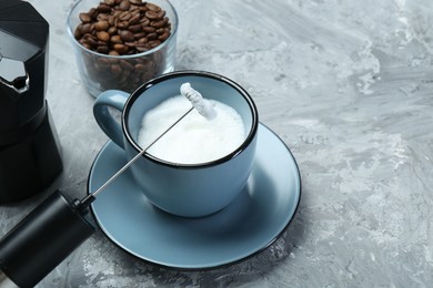 Mini mixer (milk frother), whipped milk in cup and coffee beans on grey textured table, closeup. Space for text