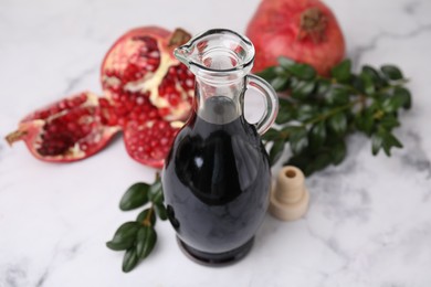 Photo of Tasty pomegranate sauce in bottle, fruits and branches on white marble table, closeup