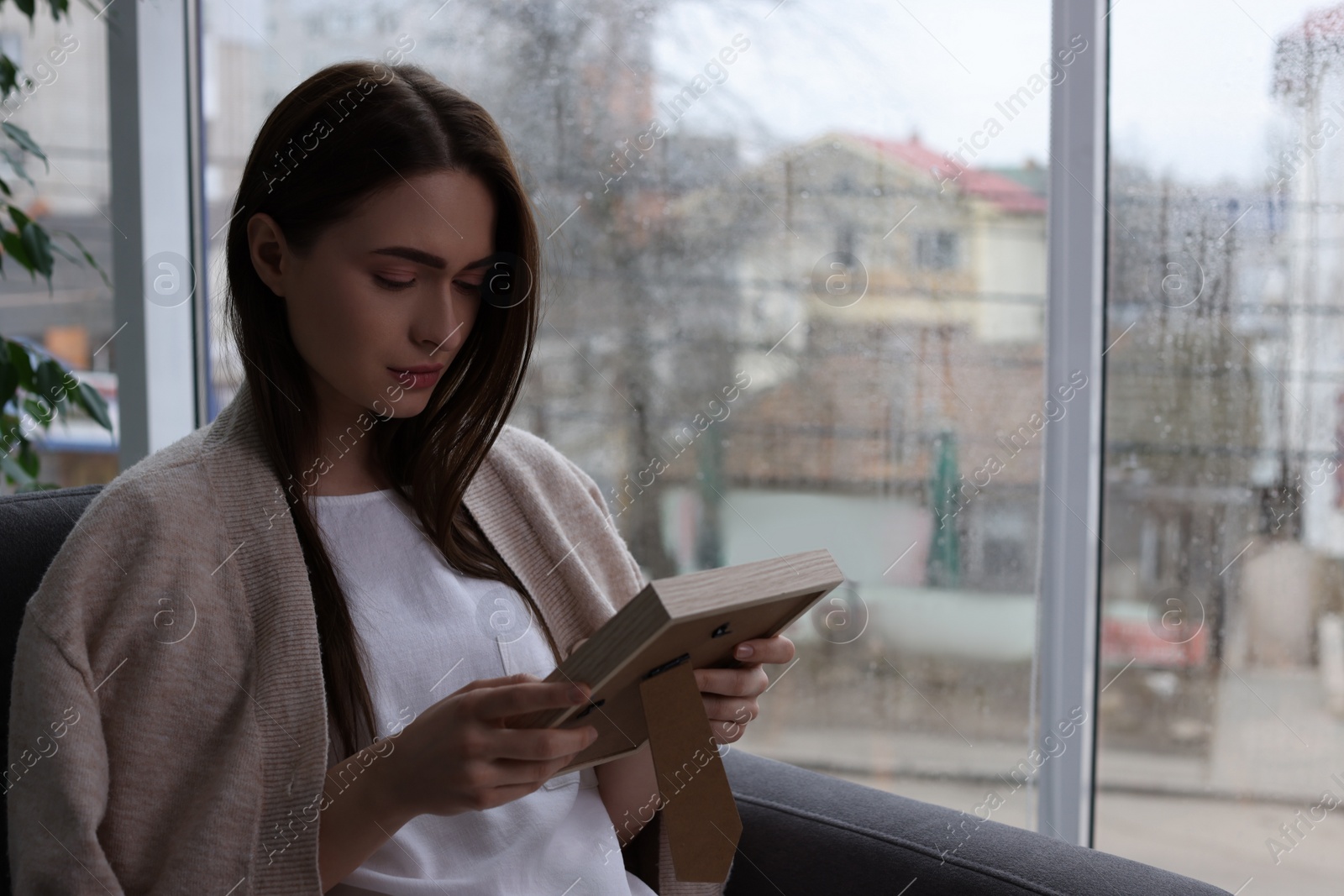 Photo of Melancholic young woman with photo frame sitting in armchair indoors, space for text. Loneliness concept