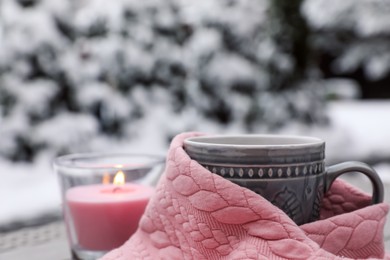 Photo of Winter morning. Cup with hot drink wrapped in pink sweater and burning candle outdoors, closeup. Space for text