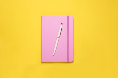 Photo of Closed pink notebook and pen on yellow background, top view