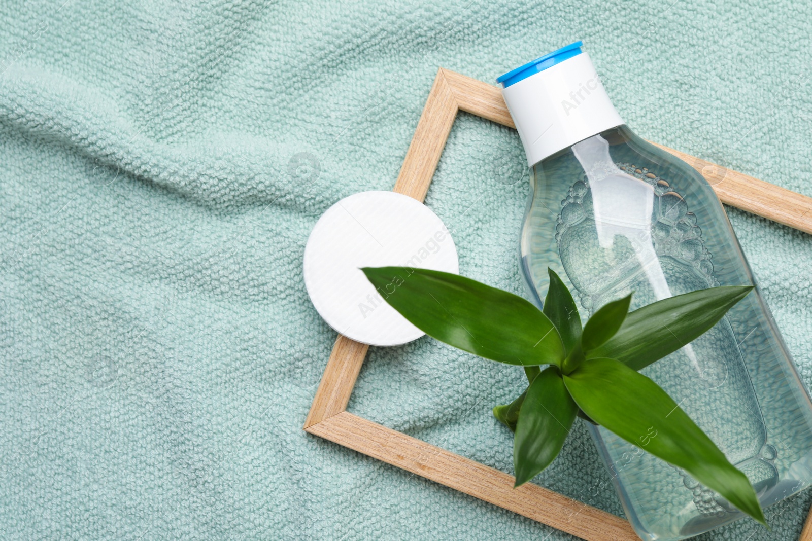 Photo of Bottle of micellar cleansing water, cotton pads and green plant on turquoise towel, flat lay. Space for text