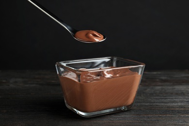 Photo of Spoon with tasty chocolate cream over glass bowl on wooden table against dark background