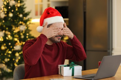 Photo of Celebrating Christmas online with exchanged by mail presents. Man in Santa hat covering eyes before opening gift box during video call on laptop at home