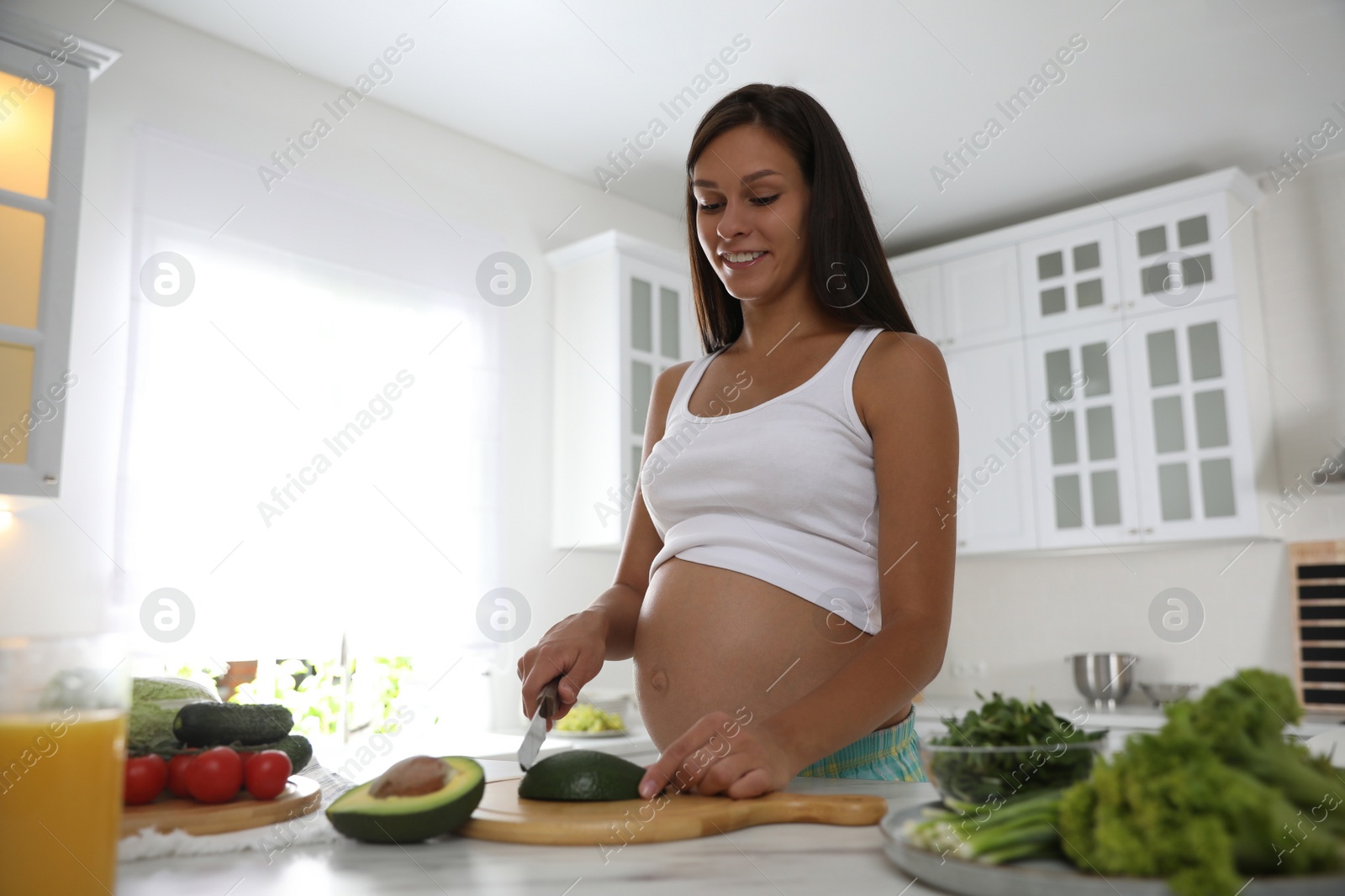 Photo of Young pregnant woman cutting avocado at table in kitchen. Taking care of baby health