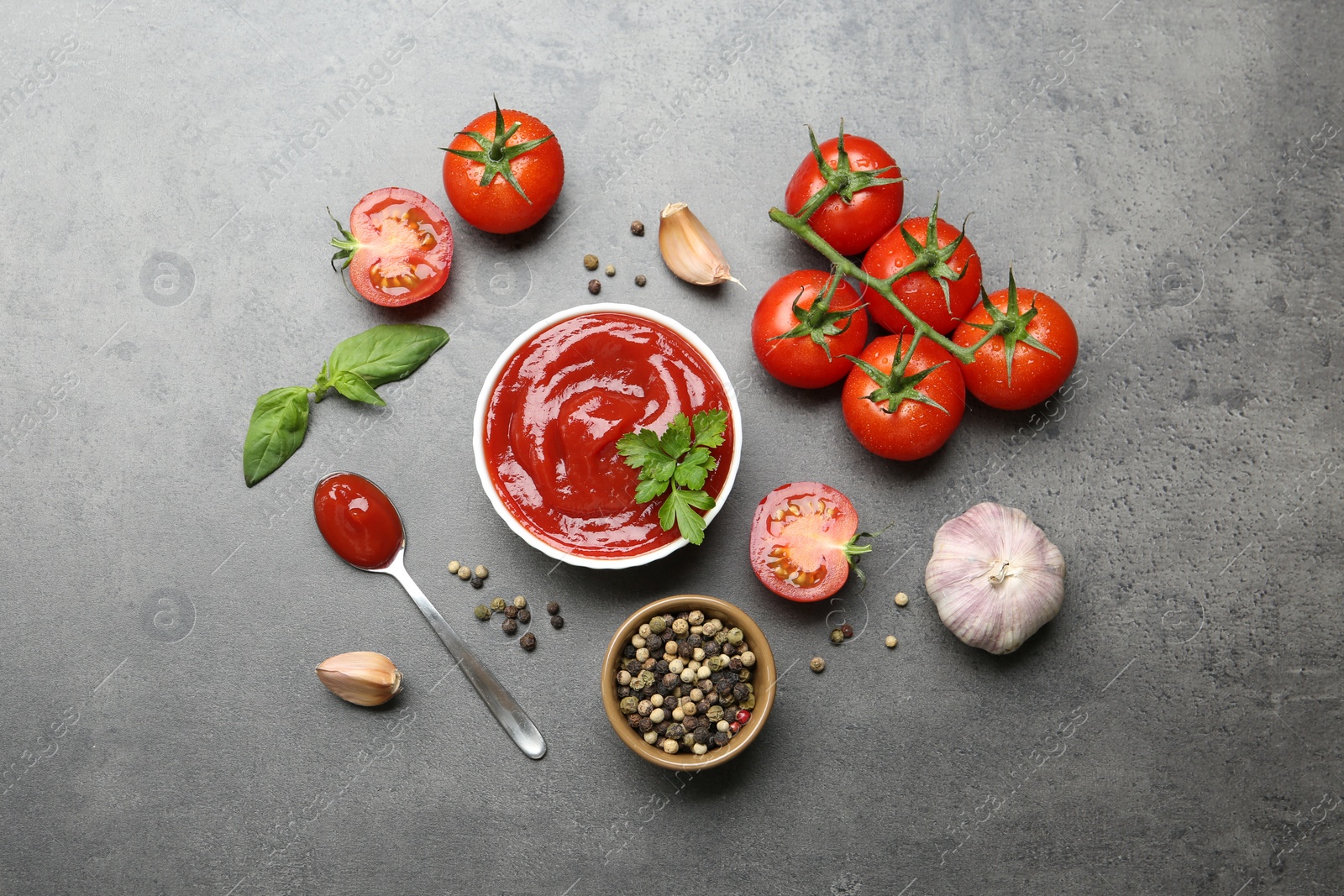 Photo of Delicious tomato ketchup in bowl, spices and products on grey textured table, flat lay