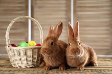 Photo of Cute bunnies and basket with Easter eggs on wooden table against blurred background