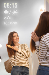 Beautiful young woman with hair brush looking at herself in smart mirror 