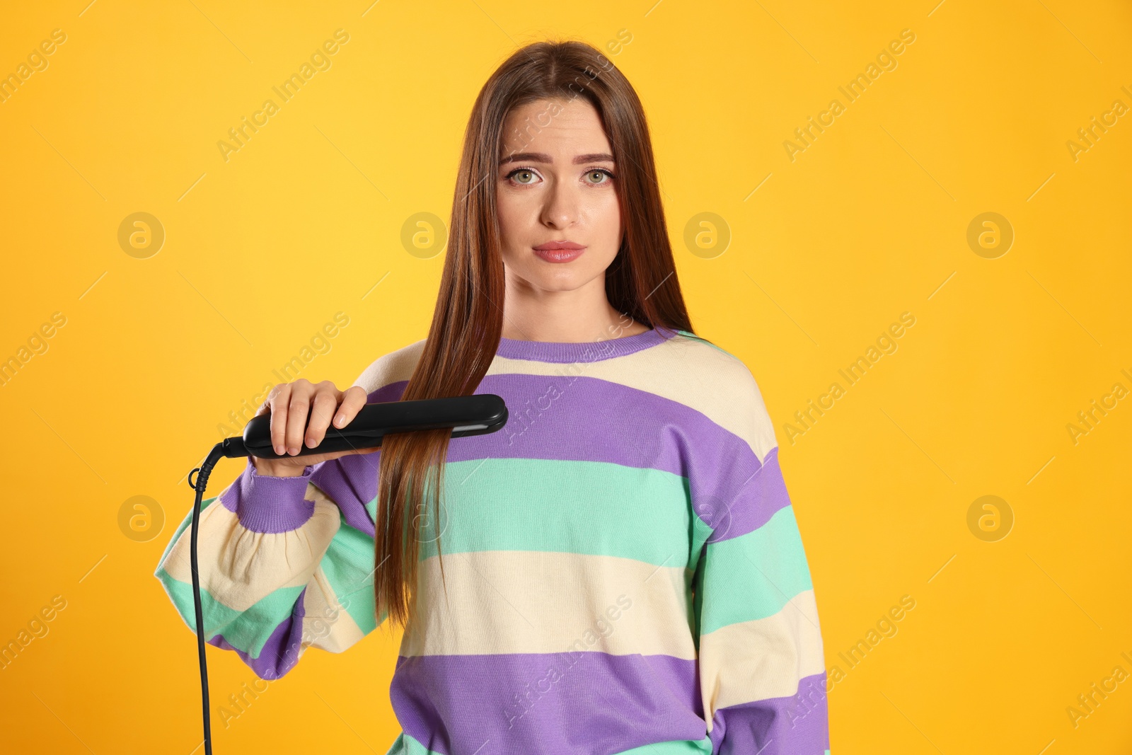 Photo of Upset young woman with flattening iron on yellow background. Hair damage