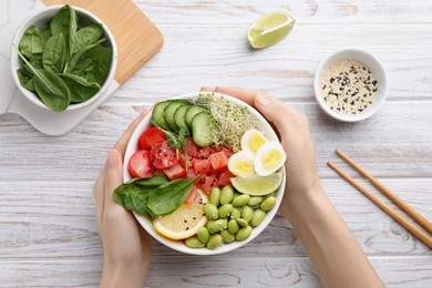 Photo of Woman holding delicious poke bowl with quail eggs, fish and edamame beans at white wooden table, top view