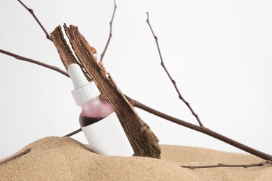 Bottle with serum, bark and branches on sand against white background, closeup. Cosmetic product