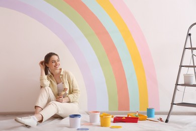 Photo of Young woman and decorator's tools near wall with painted rainbow indoors