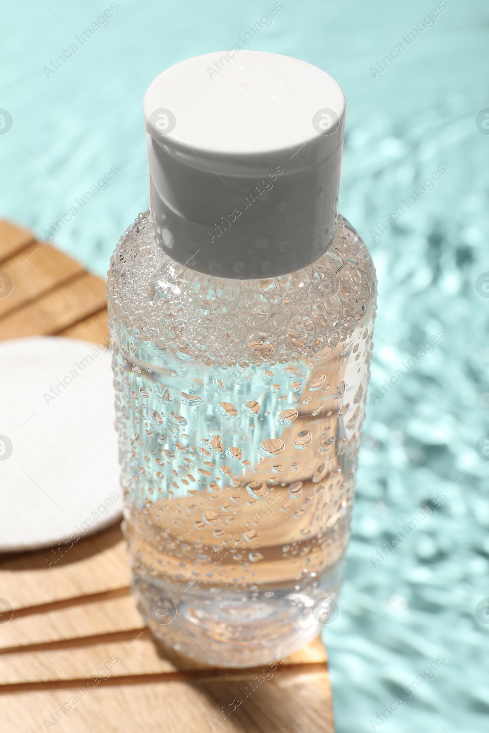 Photo of Wet bottle of micellar water and cotton pad on wooden board, closeup