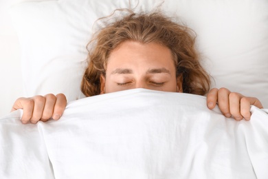 Young man covering his face with blanket while sleeping on pillow, top view. Bedtime