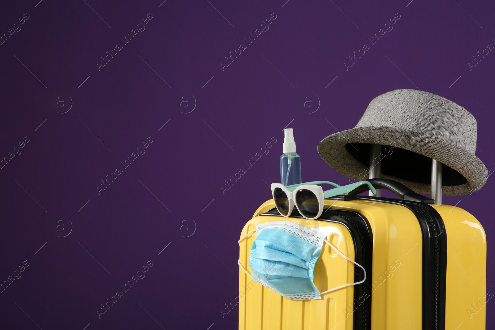 Photo of Stylish yellow suitcase with protective mask, sunglasses, hat and antiseptic spray on purple background, space for text. Travelling during coronavirus pandemic