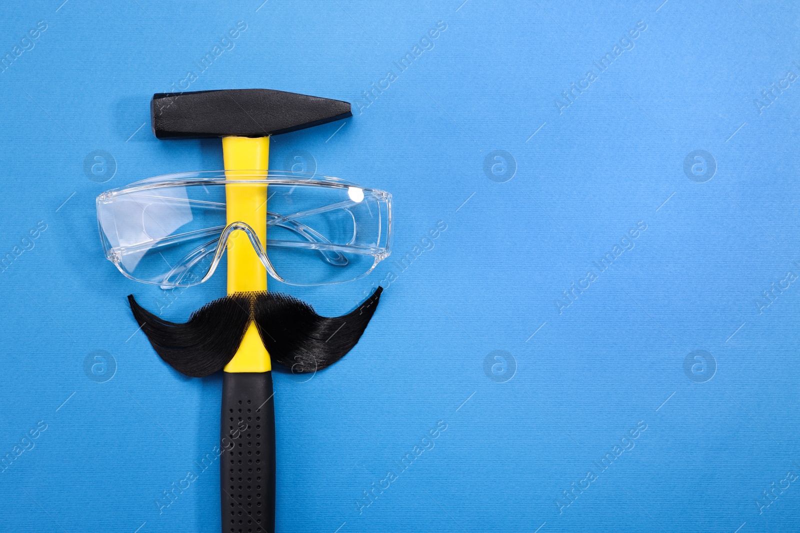 Photo of Man's face made of artificial mustache, safety glasses and hammer on blue background, top view. Space for text