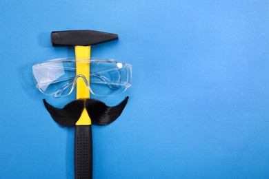 Man's face made of artificial mustache, safety glasses and hammer on blue background, top view. Space for text