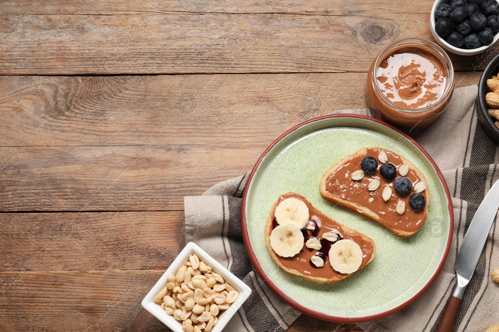 Photo of Toasts with tasty nut butter, banana slices, blueberries and peanuts on wooden table, flat lay. Space for text