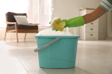 Woman holding sponge with foam over bucket indoors, closeup. Cleaning supplies