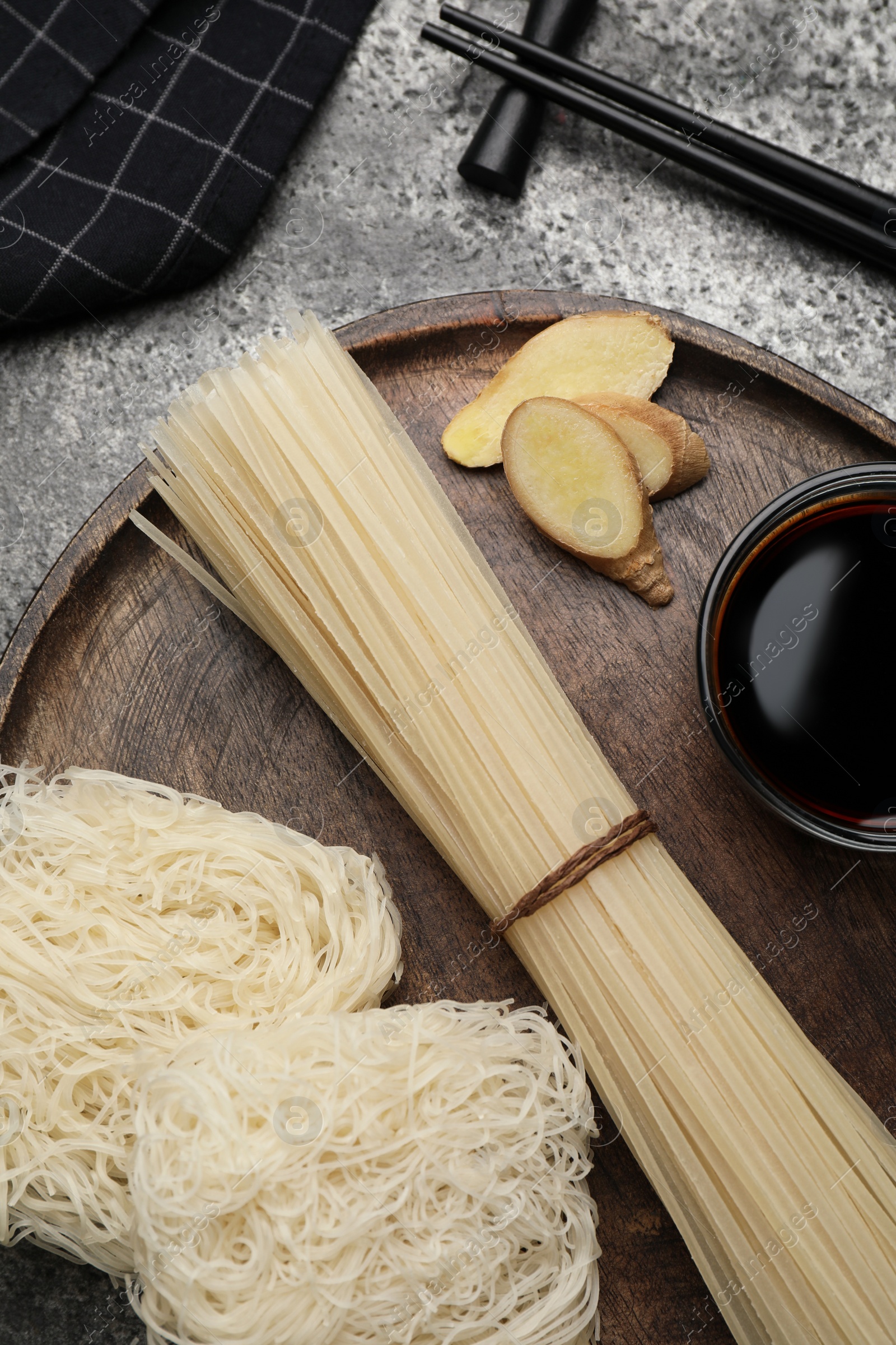 Photo of Different uncooked rice noodles, soy sauce, cut ginger and chopsticks on grey table, flat lay