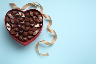 Heart shaped box with delicious chocolate candies and ribbon on light blue background, flat lay. Space for text