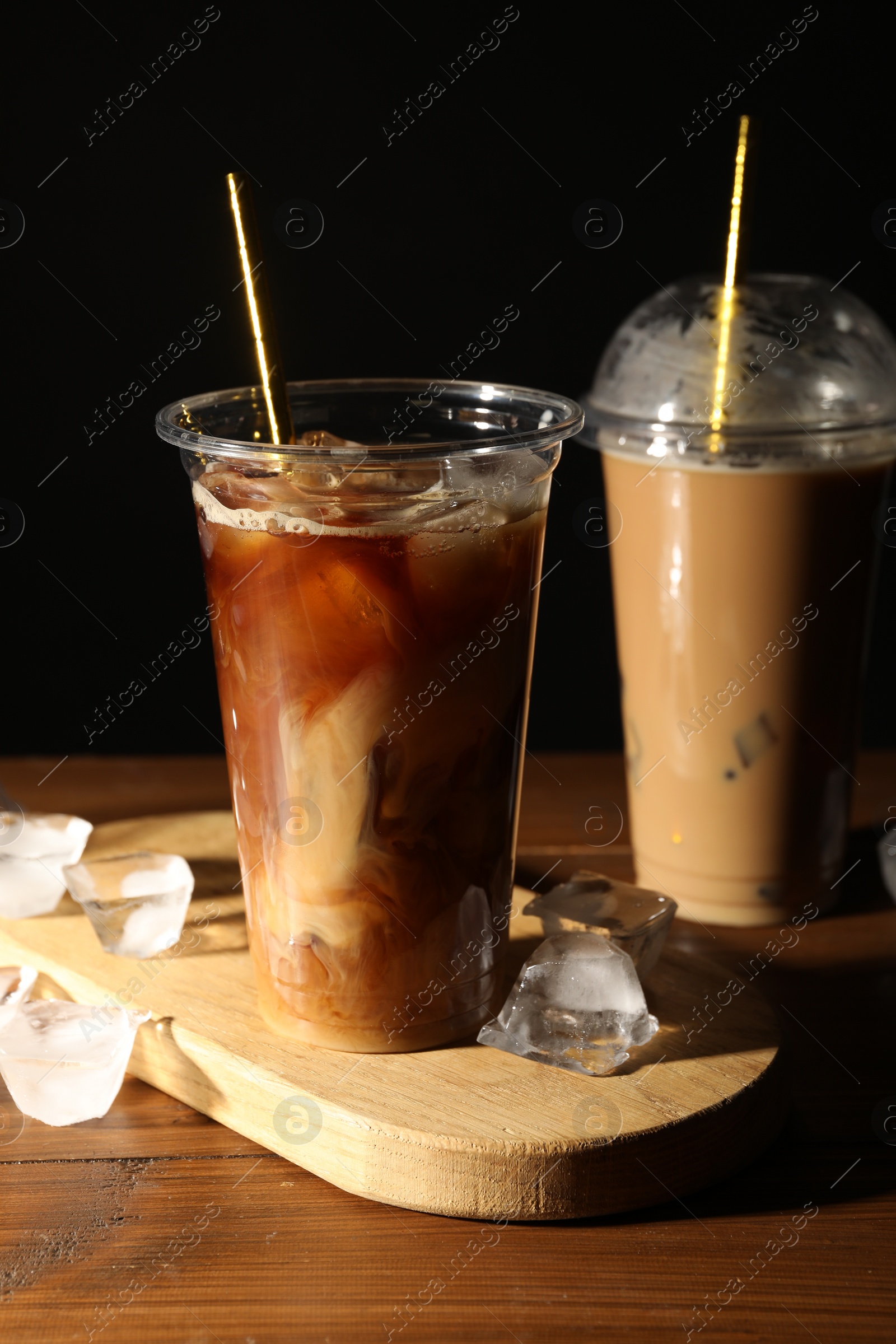 Photo of Refreshing iced coffee with milk in takeaway cups on wooden table against black background