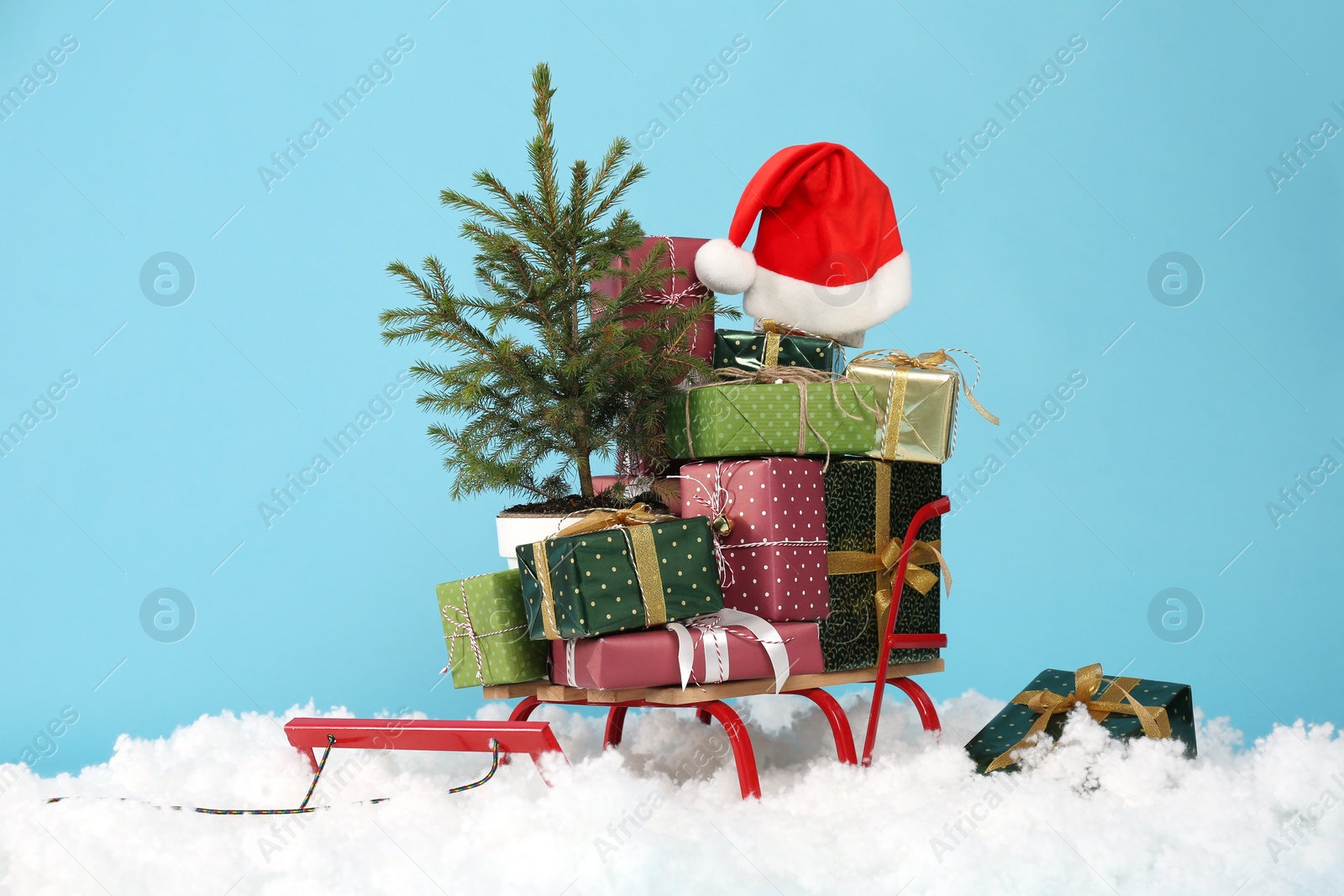 Photo of Sleigh with presents, santa hat and fir tree in artificial snow on light blue background