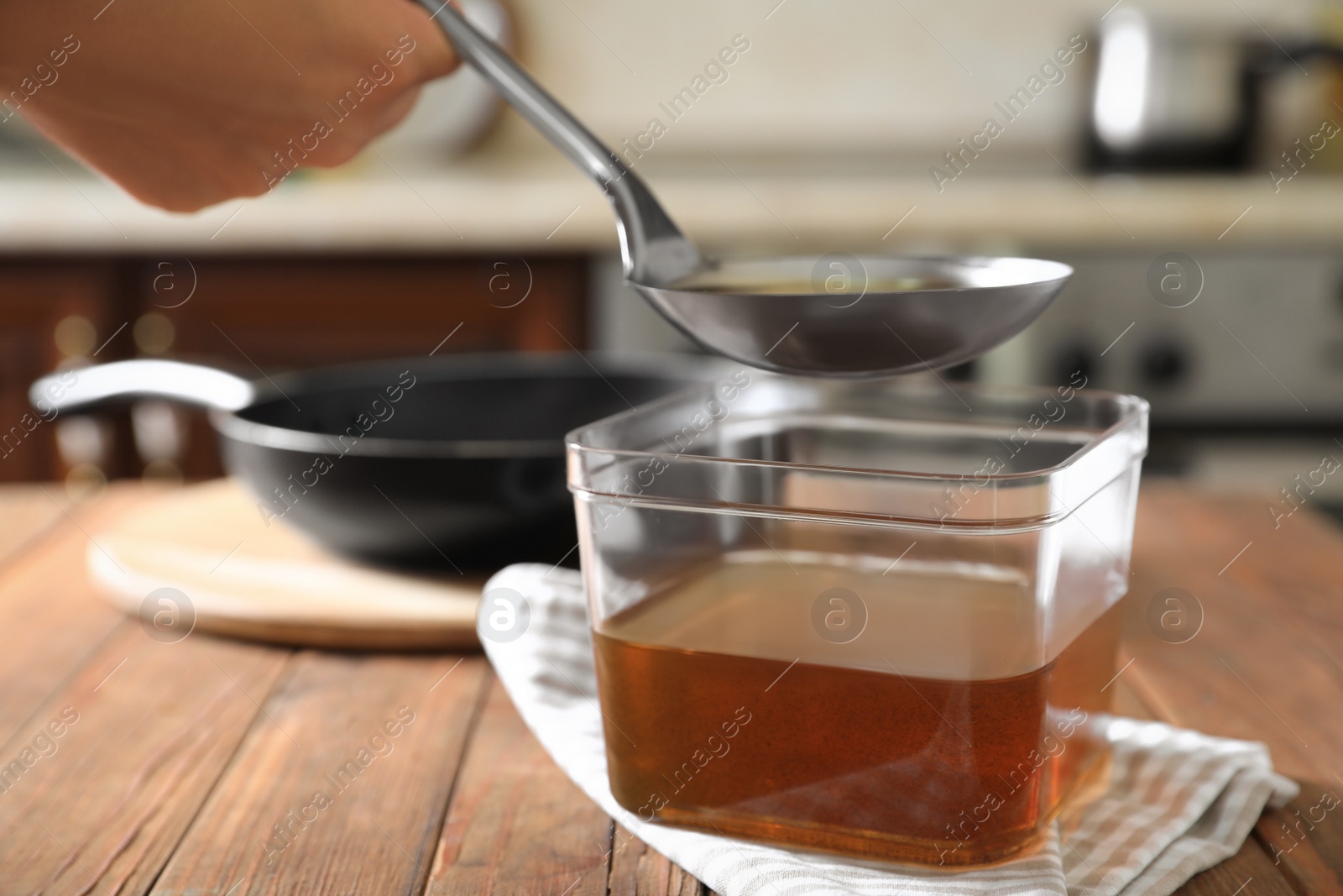 Photo of Woman pouring used cooking oil with ladle into container on wooden table in kitchen, closeup