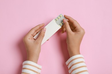 Woman holding blister with birth control pills on pink background, top view