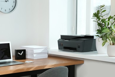 Photo of Laptop and stack of paper on wooden table near window sill with modern printer in office