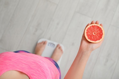 Photo of Woman holding fresh half of grapefruit while measuring her weight on floor scales, top view. Healthy diet